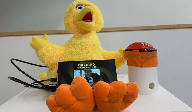 The Big Bird puppet installation with a camera, a red arcade button, and the screen reading Big Bird is Watching You